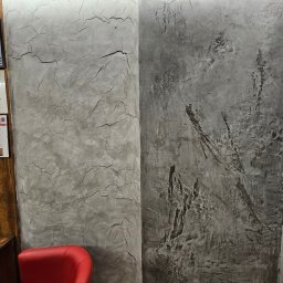 BB DESIGN - Dwa panele ścienne z dekoracyjnym tynkiem: po lewej jaśniejszy z efektem spękań, po prawej ciemniejszy z nieregularnymi wzorami, oświetlone listwą LED u góry, obok czerwone krzesło.