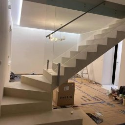 BB DESIGN - Nowoczesne, betonowe schody z szklaną balustradą w trakcie montażu, zabezpieczone kartonem, widoczna lampa sufitowa i drabina