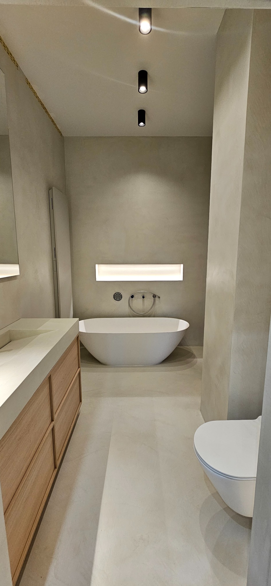 Minimalistyczna łazienka w odcieniach beżu z wolnostojącą wanną, podświetlaną wnęką nad wanną, drewnianą szafką pod umywalką i trzema punktowymi lampami sufitowymi.
