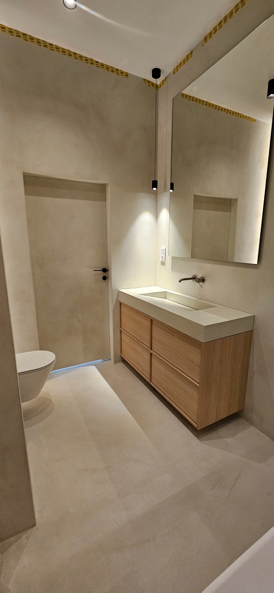 Minimalistyczna łazienka w Międzybrodziu Żywieckim z gładkimi, beżowymi ścianami, drewnianą szafką pod umywalką z betonu architektonicznego, podwieszanymi lampami i dużym lustrem z podświetleniem LED.