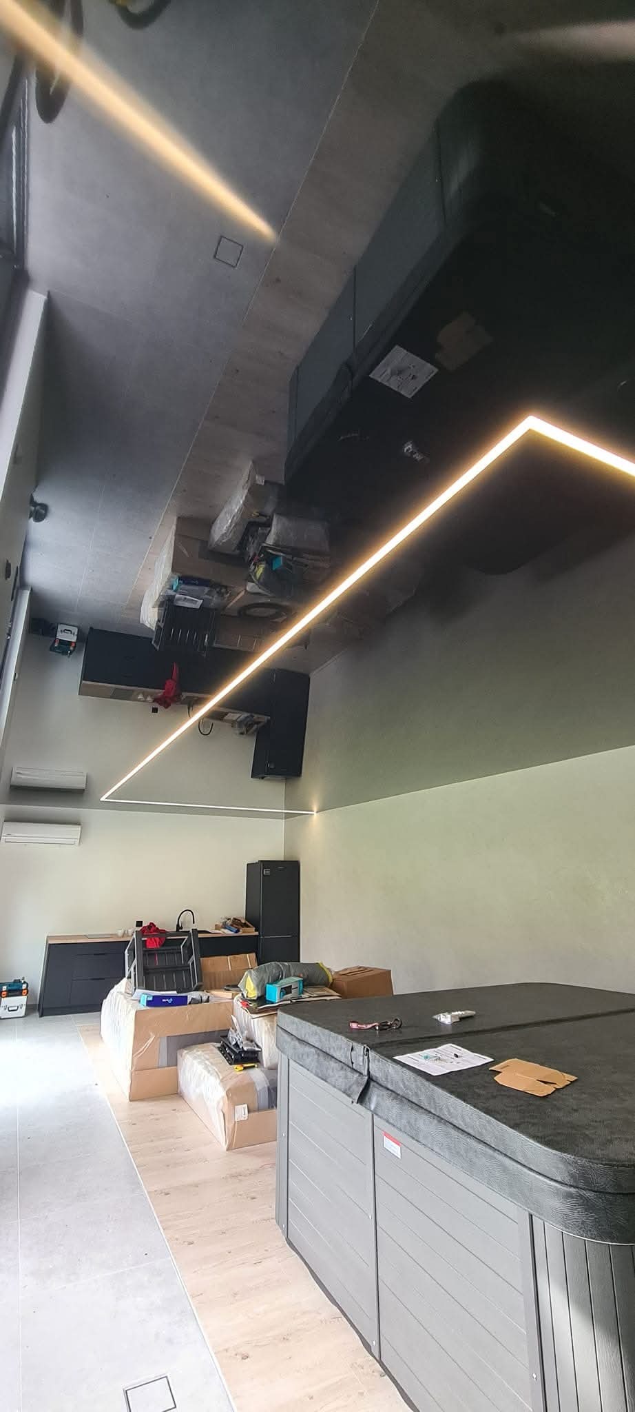 Nowoczesne wnętrze w trakcie wykańczania, z widocznym oświetleniem LED na suficie, niezapakowanym jacuzzi, meblami kuchennymi, sprzętem AGD i kartonami.