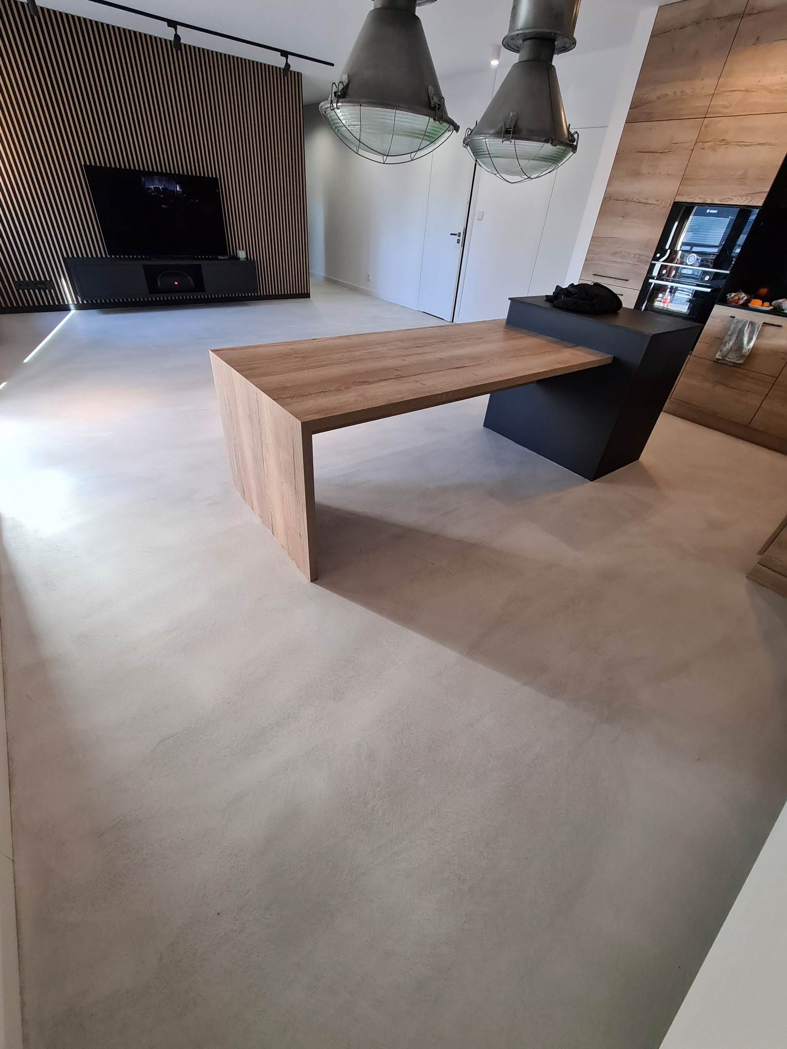 Jasnoszara, gładka posadzka z wykończeniem imitującym beton, widoczny fragment drewnianego blatu i zabudowy kuchennej, oraz dekoracyjna ściana z drewnianych listewek.