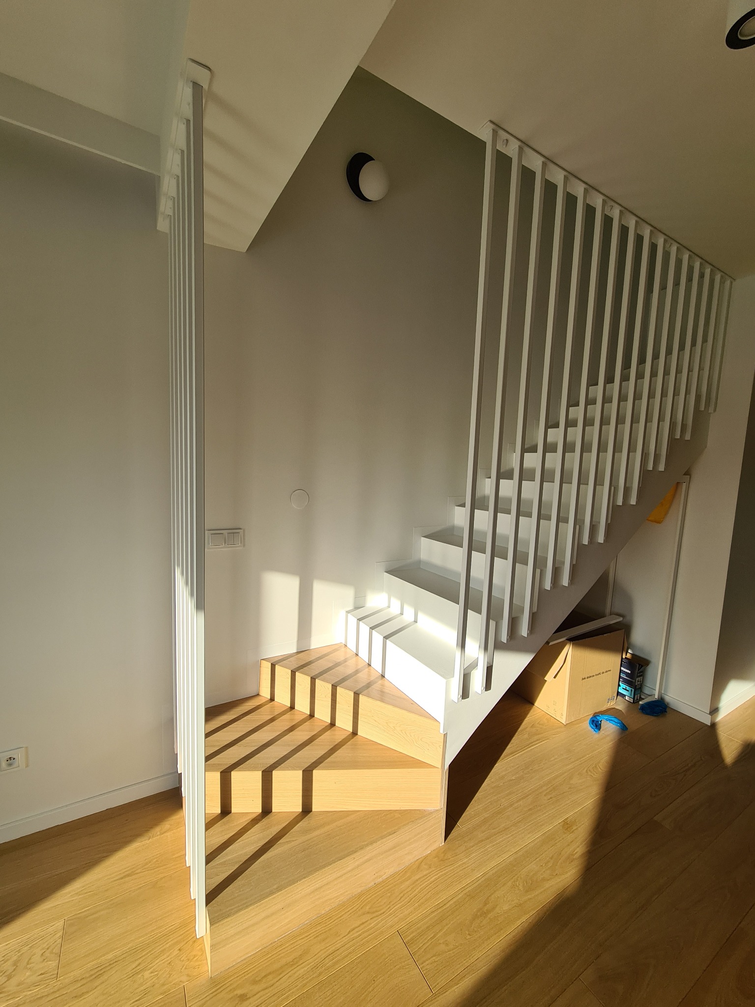 Nowoczesne, drewniane schody z białymi barierkami w jasnym wnętrzu, oświetlone promieniami słońca tworzącymi geometryczne wzory na stopniach i podłodze.