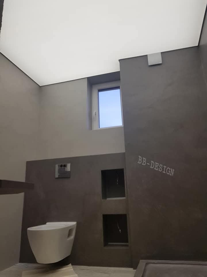 Nowoczesna łazienka z szarą, teksturowaną ścianą, białym sufitem napinanym i podwieszaną toaletą, uzupełniona o wnęki w ścianie i małe okno w górnej części ściany.