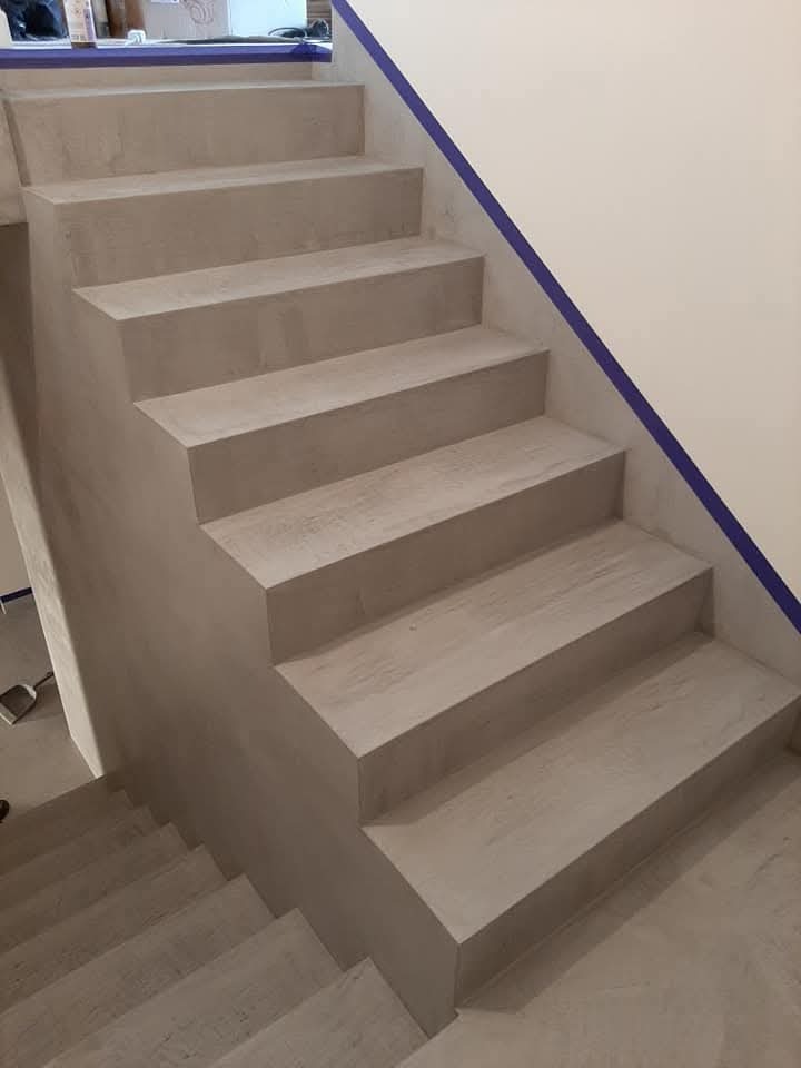 Betonowe schody wewnętrzne z widoczną strukturą powierzchni, zabezpieczone taśmą malarską przy ścianie, podczas prac wykończeniowych.