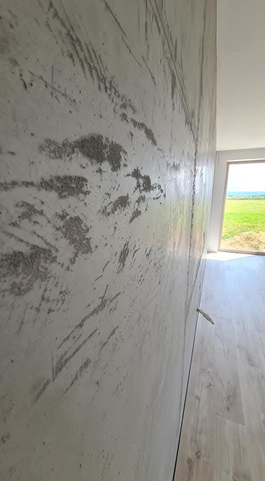 Ściana wewnętrzna z dekoracyjną, szarą fakturą imitującą beton, widok z boku, jasna podłoga i okno z widokiem na zieleń w tle.