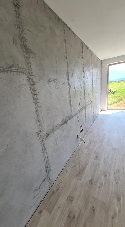 Ściana z efektem betonu architektonicznego, imitująca płyty, z widocznymi fugami, oraz jasna podłoga z paneli, widok na zielony krajobraz przez okno.