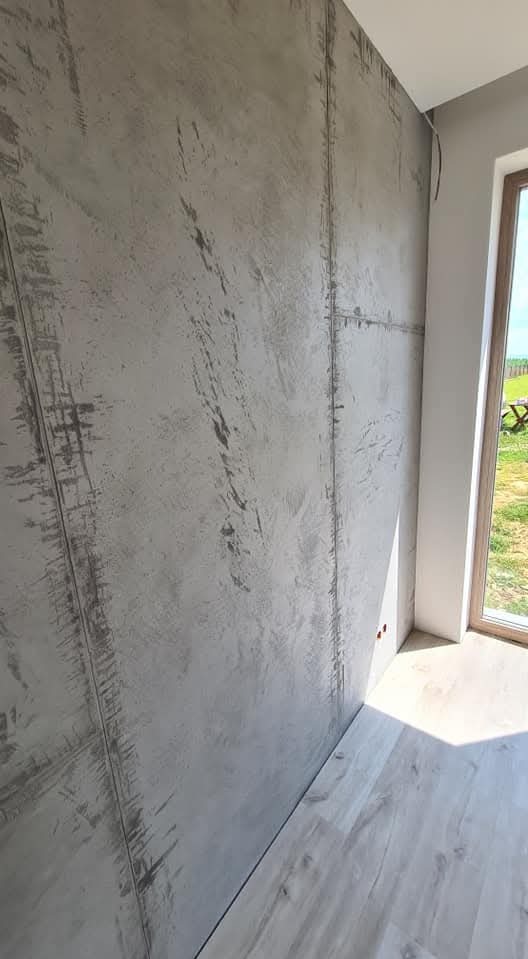 Ściana z szarą, dekoracyjną fakturą imitującą beton, podzielona pionowymi i poziomymi liniami, z widoczną podłogą z paneli i oknem z widokiem na zieleń.