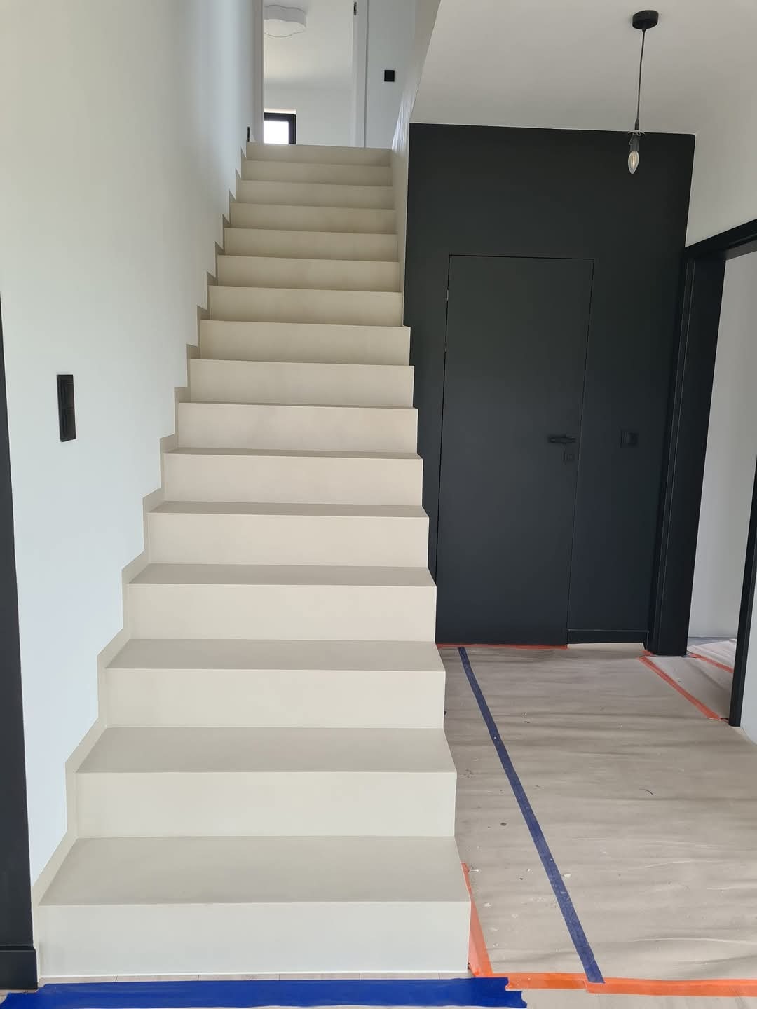 Nowoczesne, minimalistyczne schody o jasnobeżowym odcieniu, prowadzące na wyższą kondygnację, z czarną ścianą i drzwiami w tle, zabezpieczone folią malarską na podłodze.