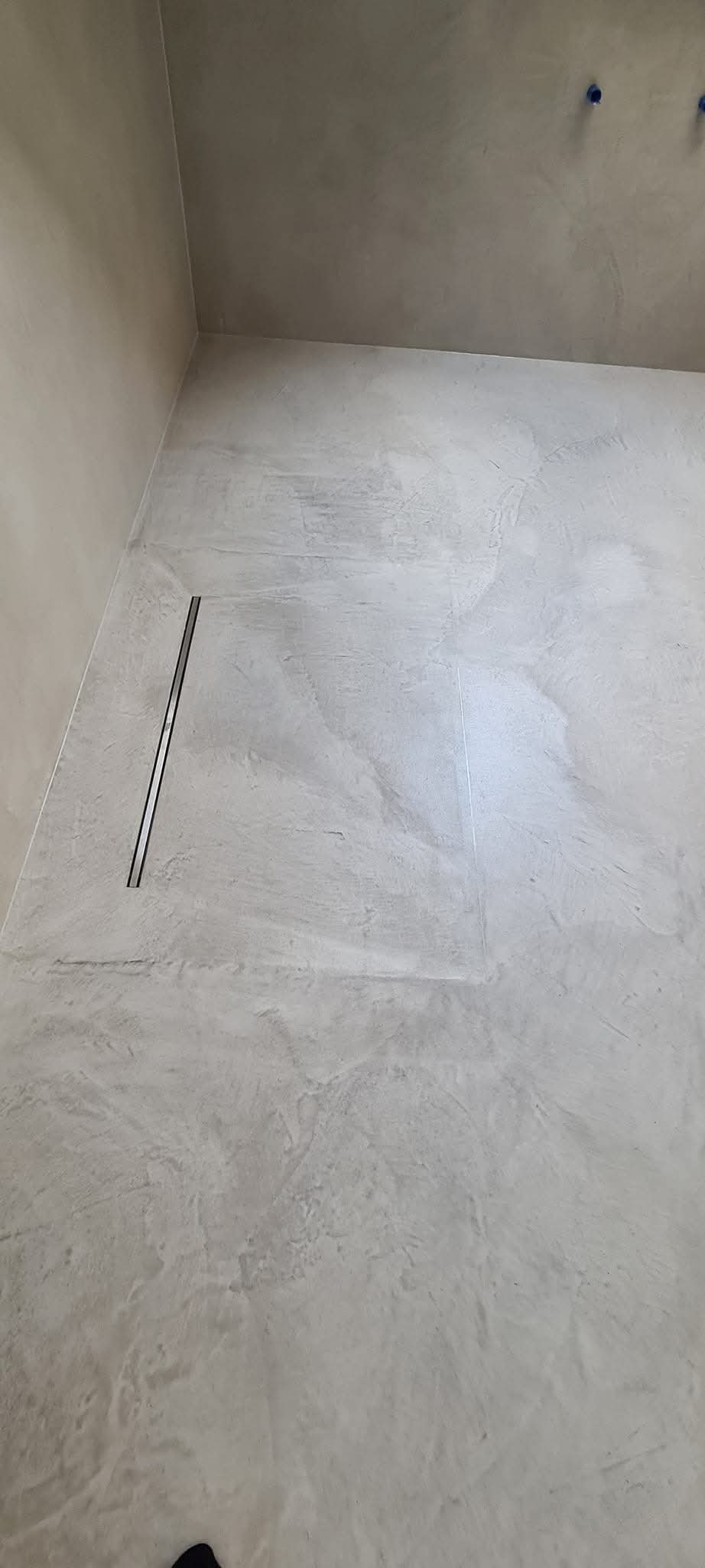 Szara posadzka żywiczna w łazience z odpływem liniowym przy ścianie, widoczne niebieskie zaślepki wystających rur na ścianie.