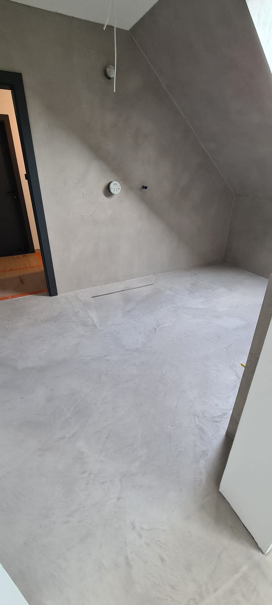 Szara, gładka podłoga w pokoju na poddaszu z widoczną kratką ściekową w posadzce i czarną ościeżnicą drzwiową.