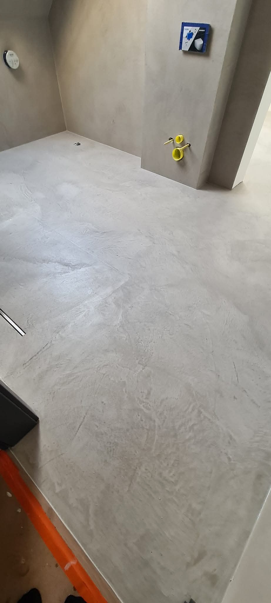 Szara, gładka posadzka w łazience z widocznymi przyłączami hydraulicznymi i odpływem liniowym.