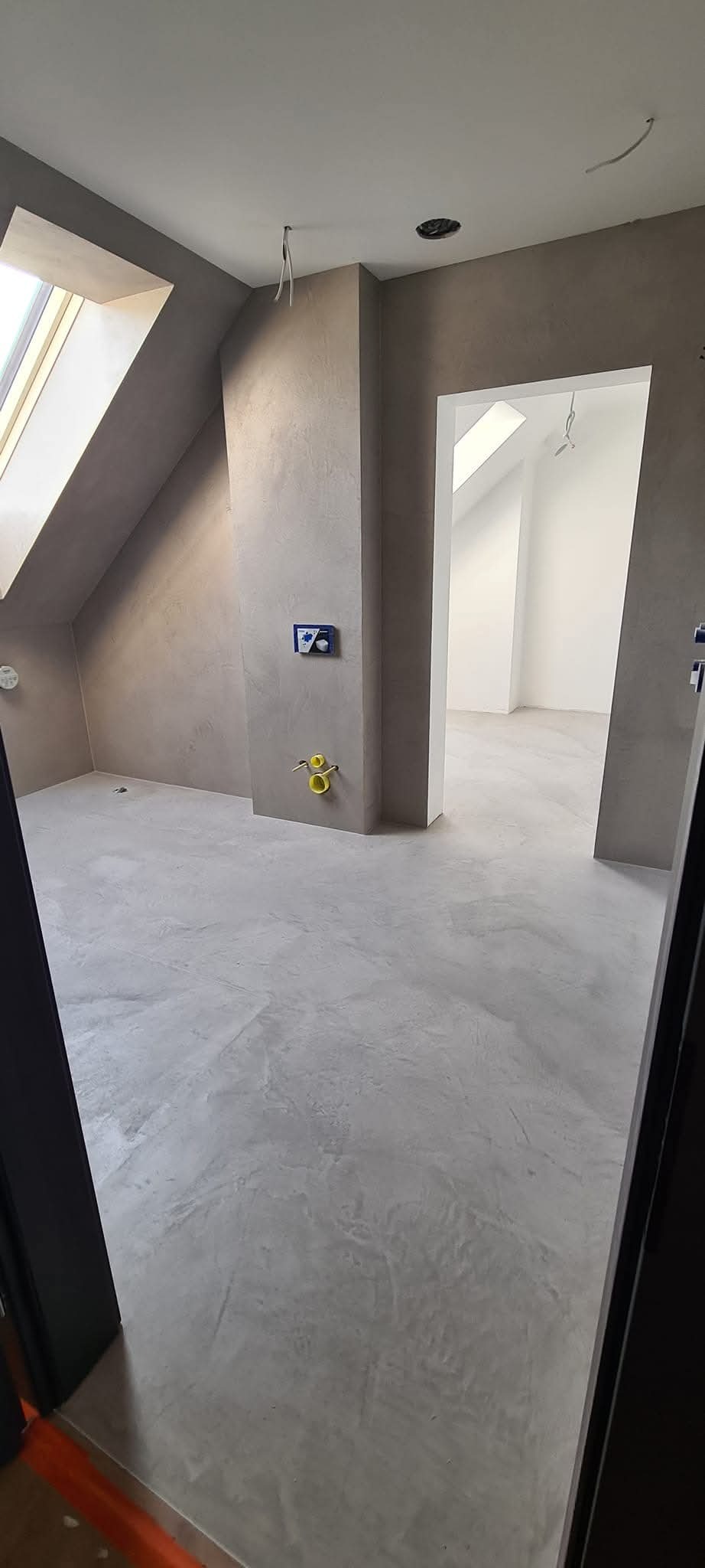 Szare wnętrze poddasza z wykończoną podłogą, widoczne przyłącza hydrauliczne i elektryczne, okno dachowe w skosie.