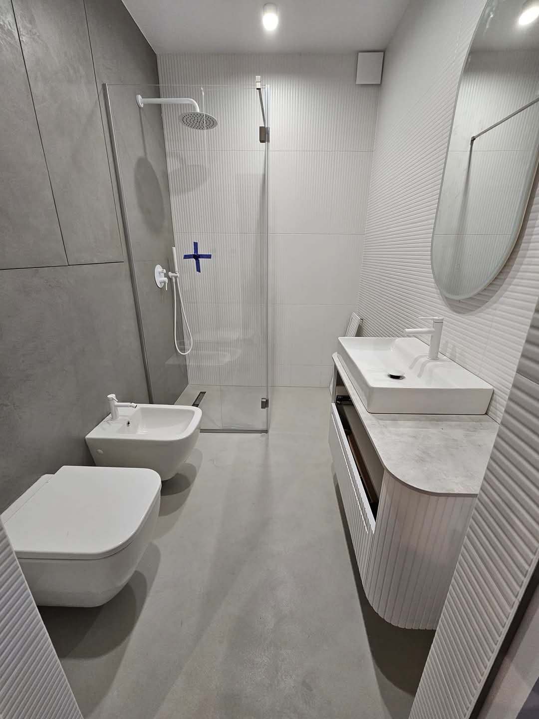 Nowoczesna łazienka z kabiną prysznicową typu walk-in, podwieszaną toaletą i bidetem, umywalką nablatową na szafce z ryflowanym frontem oraz dużym owalnym lustrem. Szare ściany z betonową fakturą...