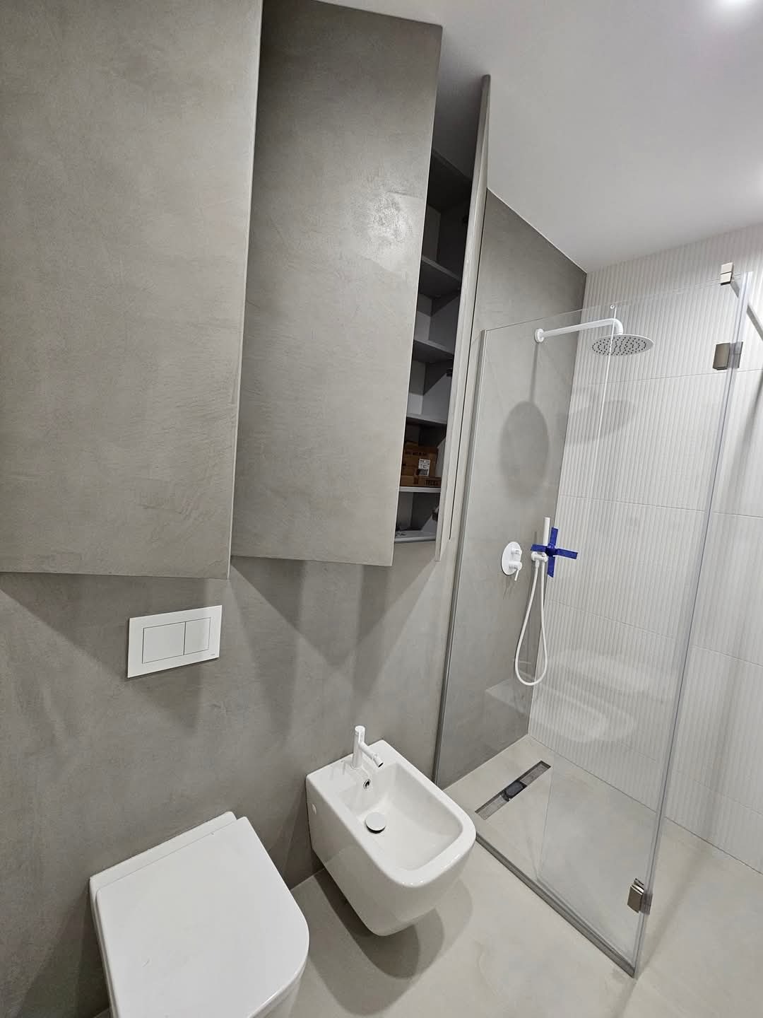 Minimalistyczna łazienka w odcieniach szarości z otwartą szafką w zabudowie, wiszącą toaletą, bidetem i kabiną prysznicową typu walk-in z odpływem liniowym.