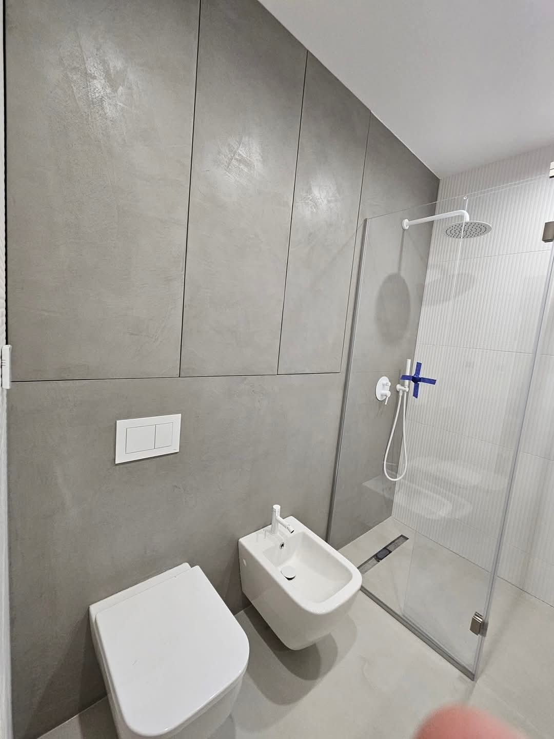 Nowoczesna łazienka z szarą ścianą o fakturze betonu, białą armaturą (toaleta i bidet) oraz kabiną prysznicową ze szklanymi drzwiami.