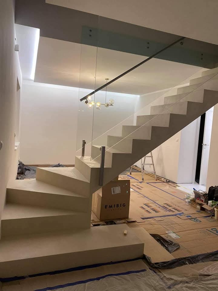 Nowoczesne, betonowe schody z szklaną balustradą w trakcie montażu, zabezpieczone kartonem, widoczna lampa sufitowa i drabina