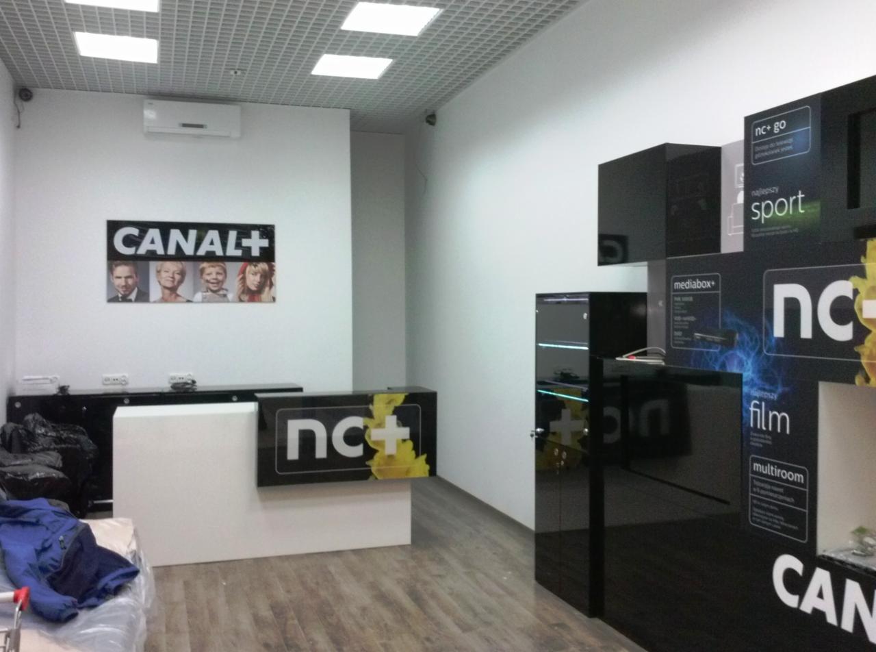 Wnętrze punktu obsługi klienta z logo Canal+ i nc+, widoczne elementy wyposażenia takie jak lada recepcyjna, regały z ekspozycją produktów, oświetlenie sufitowe i reklamy wizualne.