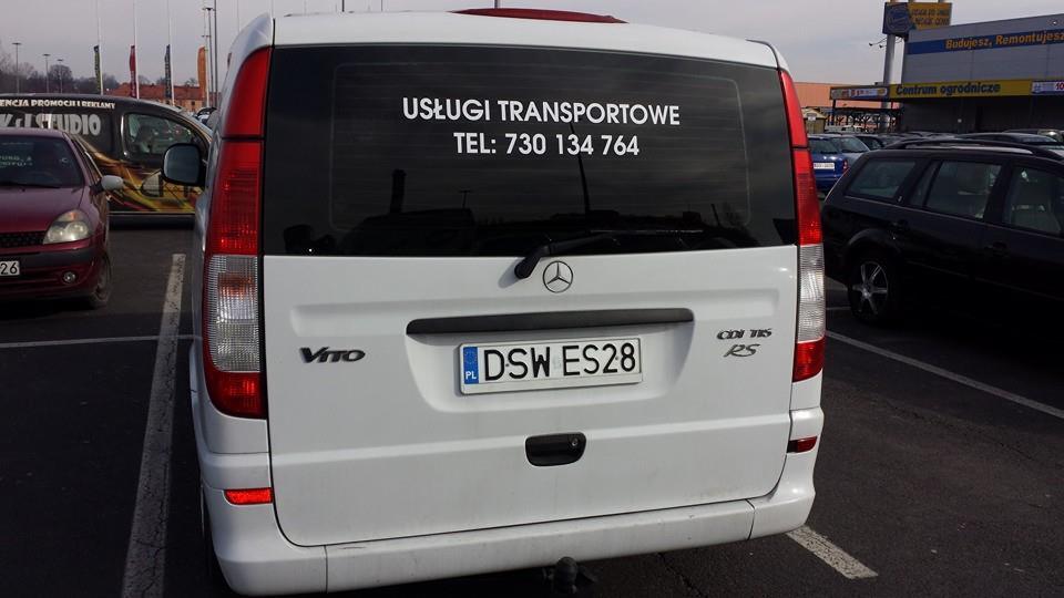 Tył białego busa Mercedes Vito z napisem 'Usługi Transportowe, Tel: 730 134 764' na tylnej szybie, zaparkowany na parkingu przed centrum handlowym.