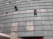 Dwaj alpiniści myją szklaną fasadę wysokiego biurowca, zawieszeni na linach. Widoczne odbicia w oknach i fragment budynku na dole. Ujęcie z dołu.