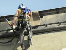 Pracownik w uprzęży, z linami i narzędziami, wykonuje prace na wysokości na tle jasnego nieba. Widok z dołu, skupienie na bezpieczeństwie i profesjonalizmie.