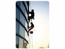 Dwóch alpinistów w uprzężach, na linie, myje szklaną fasadę budynku w słoneczny dzień. Widok z dołu, sylwetki na tle jasnego nieba.