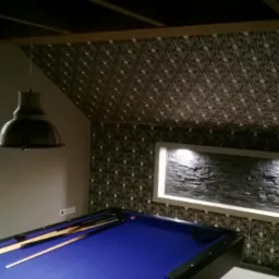 Stylowe wykończenie pokoju bilardowego z akcentem kamiennej ściany, wzorzystej tapety sufitowej i designerskiej lampy nad niebieskim stołem bilardowym.