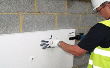 Pracownik w kasku i kamizelce odblaskowej wierci otwór w białej płycie izolacyjnej przymocowanej do ściany z szarego betonu komórkowego. Widoczna wiertarka udarowa i rękawica robocza.