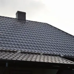 Ciemny, metalowy dach z dwoma kominami z cegły. Widoczne rynny i elementy wykończeniowe dachu. Dachówka ułożona równo, tworzy regularny wzór.
