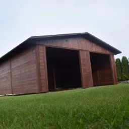 Drewniana wiata garażowa z dwoma otwartymi wjazdami, na trawniku, z przyczepką w tle. Widoczny fragment domu i ogrodzenie.