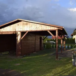 Drewniana wiata z zadaszeniem, widoczna z boku, z elementami konstrukcyjnymi w kolorze ciemnego brązu, na tle domu i pochmurnego nieba.