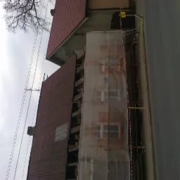 Budynek w trakcie remontu elewacji, zabezpieczony siatką ochronną. Widoczny fragment dachu z blachy trapezowej. Ujęcie z dołu, pochmurne niebo w tle.