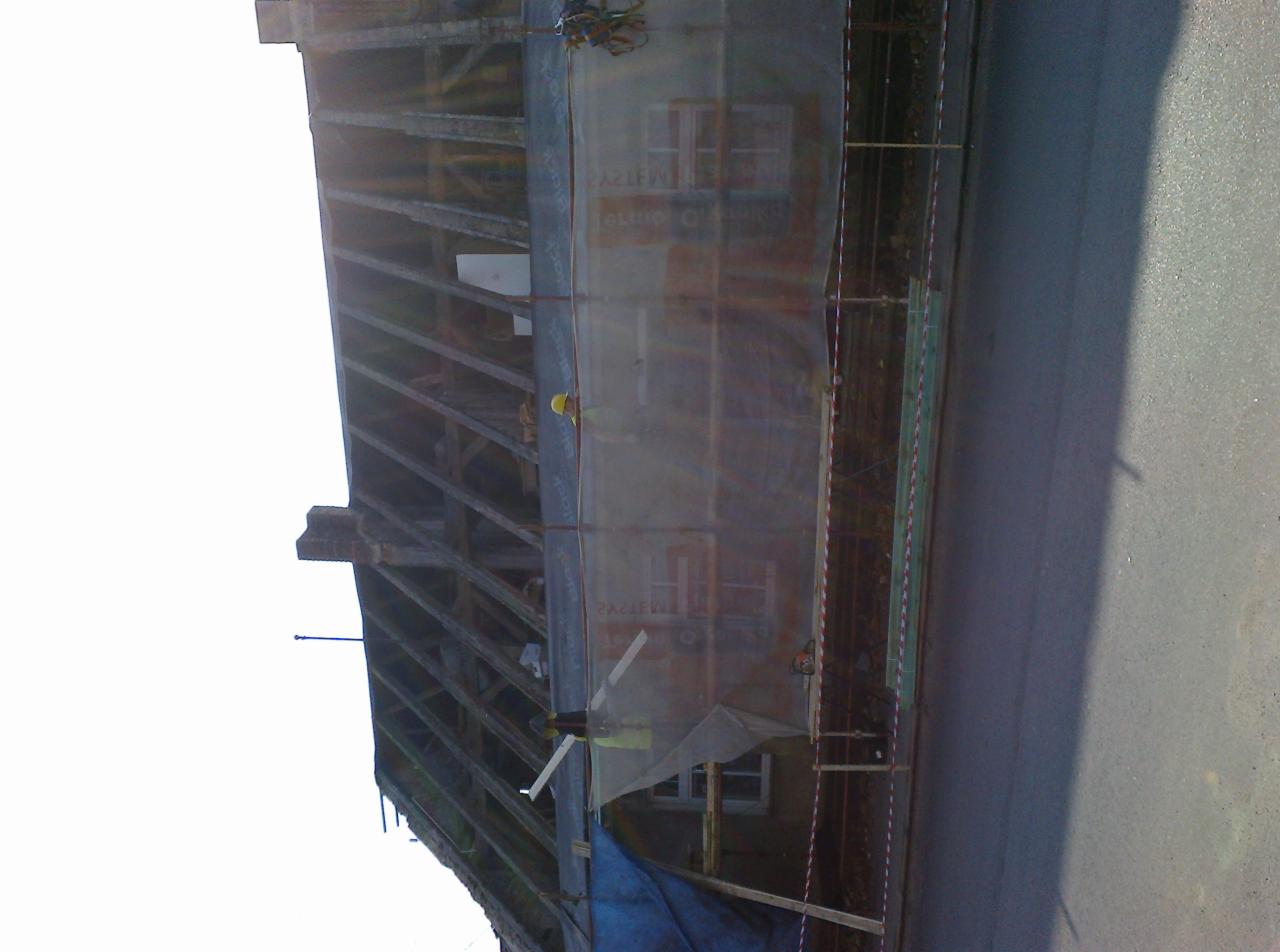 Konstrukcja drewniana dachu w trakcie prac, zabezpieczona siatką ochronną, widoczny pracownik w kasku. Ujęcie z dołu, fragment ulicy w kadrze.