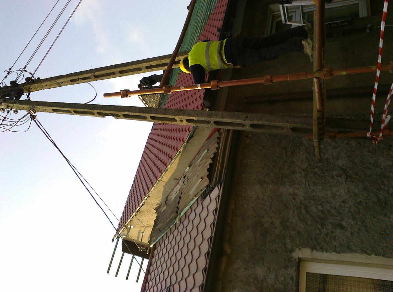 Pracownik w żółtej kamizelce na rusztowaniu przy dachu z czerwonej dachówki, wykonujący prace dekarskie. Widoczny słup elektryczny i fragment elewacji budynku.