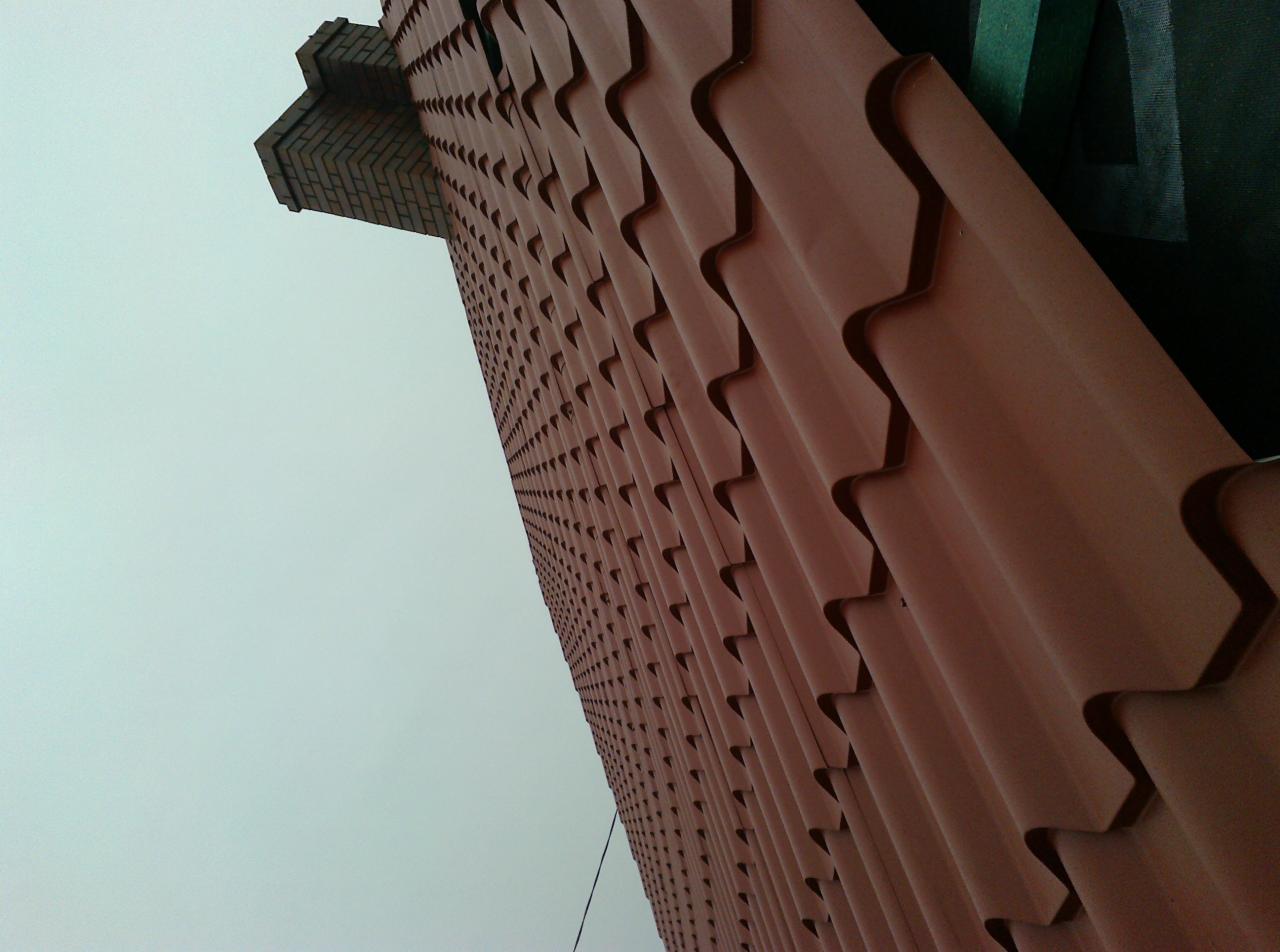 Czerwona dachówka ceramiczna na ukośnej połaci dachu, widziana z dołu, z fragmentem komina w lewym górnym rogu na tle szarego nieba.