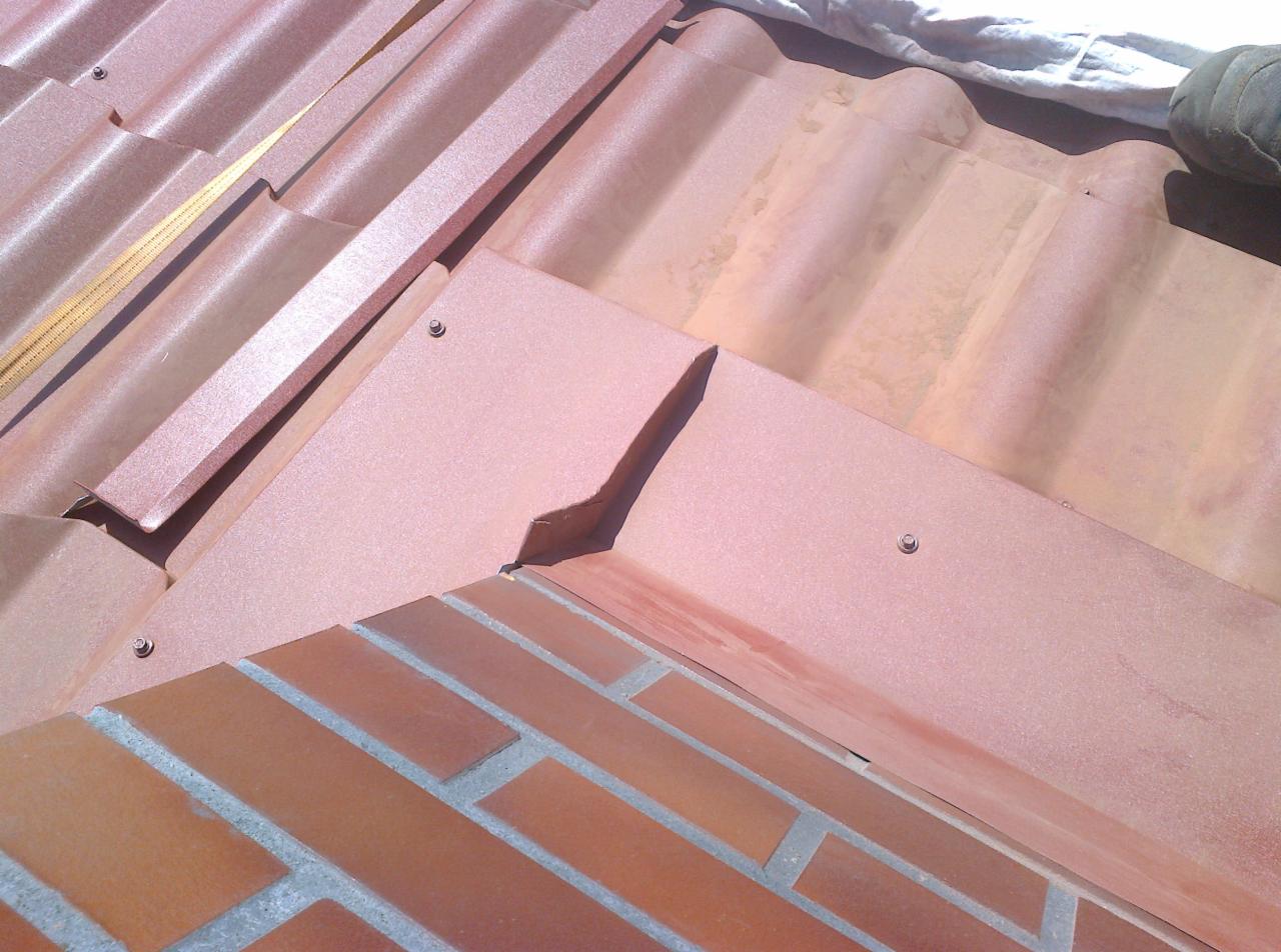 Szczegółowe ujęcie fragmentu dachu z ceglastą dachówką karpiówką i obróbką blacharską w kolorze zbliżonym do dachówki, widoczny mur z cegły.