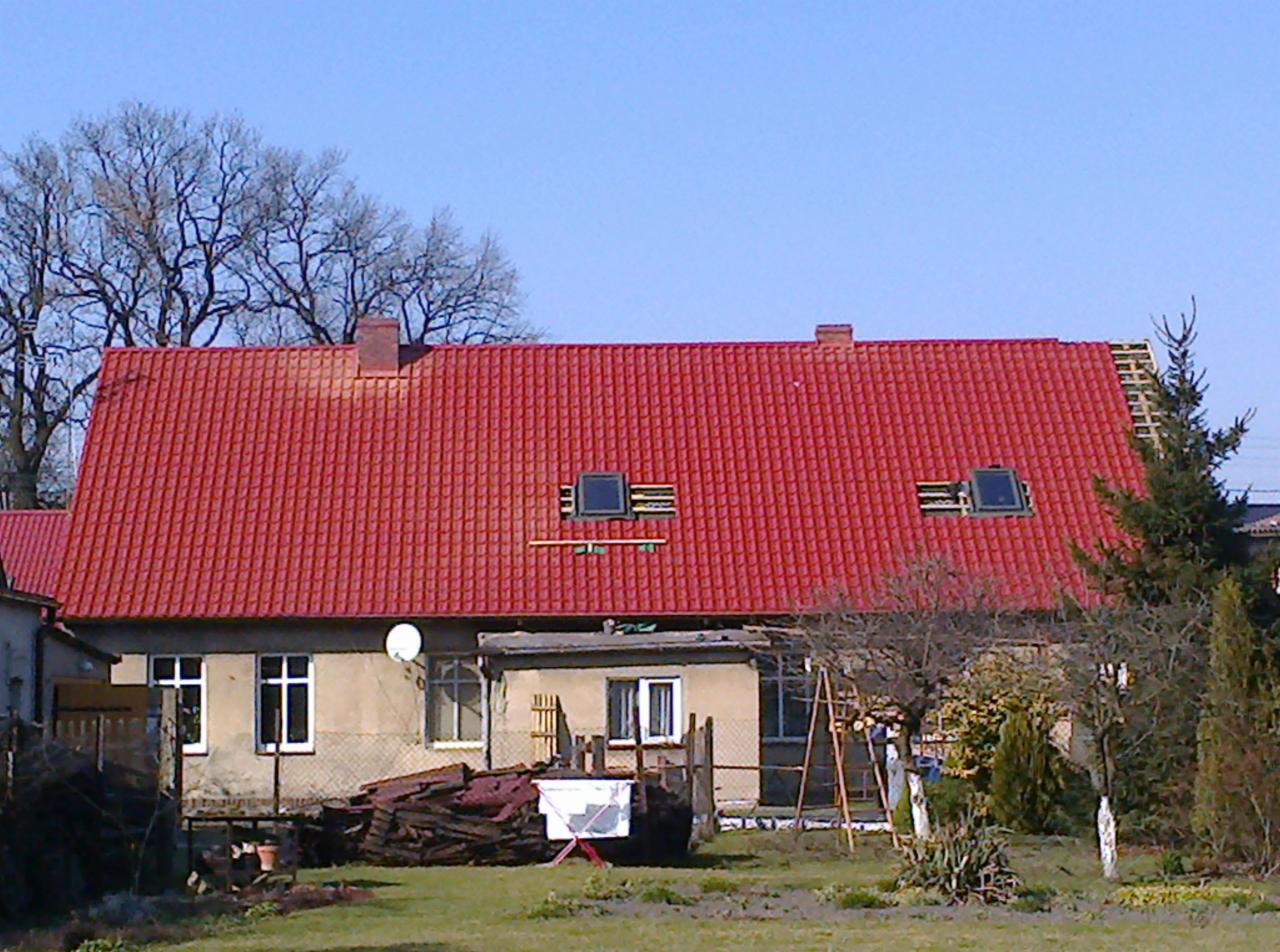 Dom z czerwonym dachem krytym dachówką, widoczne okna dachowe i komin. Część dachu w trakcie prac wykończeniowych, widoczne elementy konstrukcyjne.