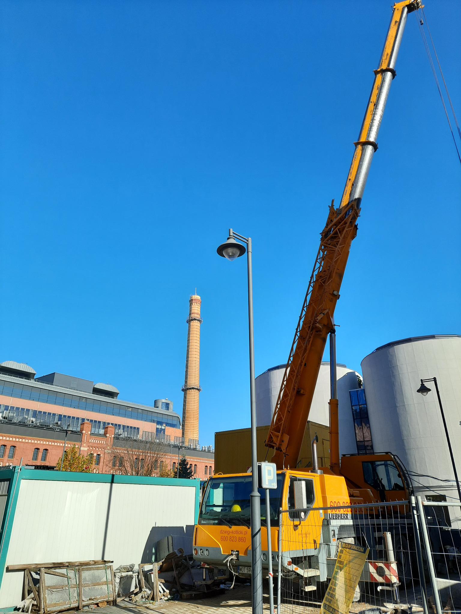 Żółty dźwig samochodowy Liebherr z wysięgnikiem na tle błękitnego nieba i ceglanego komina, widoczny fragment placu budowy z kontenerami i ogrodzeniem.