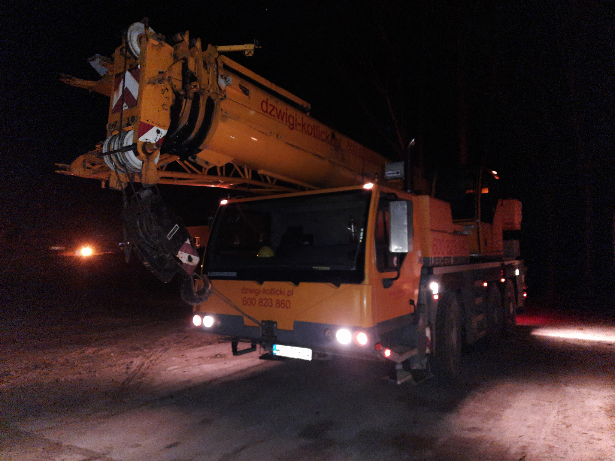 Żółty dźwig samochodowy Liebherr z wysięgnikiem, widoczny w nocy z włączonymi światłami, na ziemi ślady opon, w tle ciemność i słabe światło.