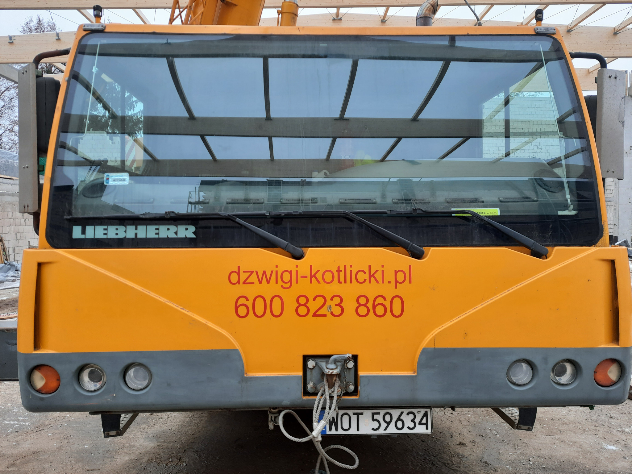 Żółty dźwig Liebherr z widocznym numerem telefonu i adresem strony internetowej na kabinie, zaparkowany na placu budowy, widok z tyłu.