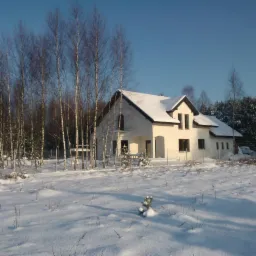 Biały dom jednorodzinny z ciemnym dachem, pokryty śniegiem, otoczony drzewami bez liści w zimowej scenerii pod błękitnym niebem.