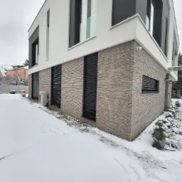 Nowoczesny dom z cegły klinkierowej z czarnymi zewnętrznymi roletami w zimowej scenerii, śnieg pokrywa trawnik i krzewy.