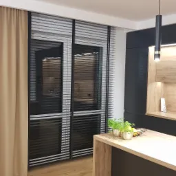 Nowoczesna kuchnia w Otwocku z oknami przesłoniętymi czarnymi żaluzjami poziomymi, widok z wnętrza, ciepłe oświetlenie podkreśla drewniane elementy zabudowy.