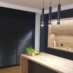 Nowoczesna kuchnia z drewnianymi szafkami, czarnymi poziomymi listwami okiennymi i beżową zasłoną po lewej stronie, oświetlona trzema wiszącymi lampami.