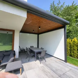 Nowoczesna pergola tarasowa z drewnianym sufitem i białymi ścianami bocznymi, wyposażona w szary stół i krzesła, otoczona zielenią.