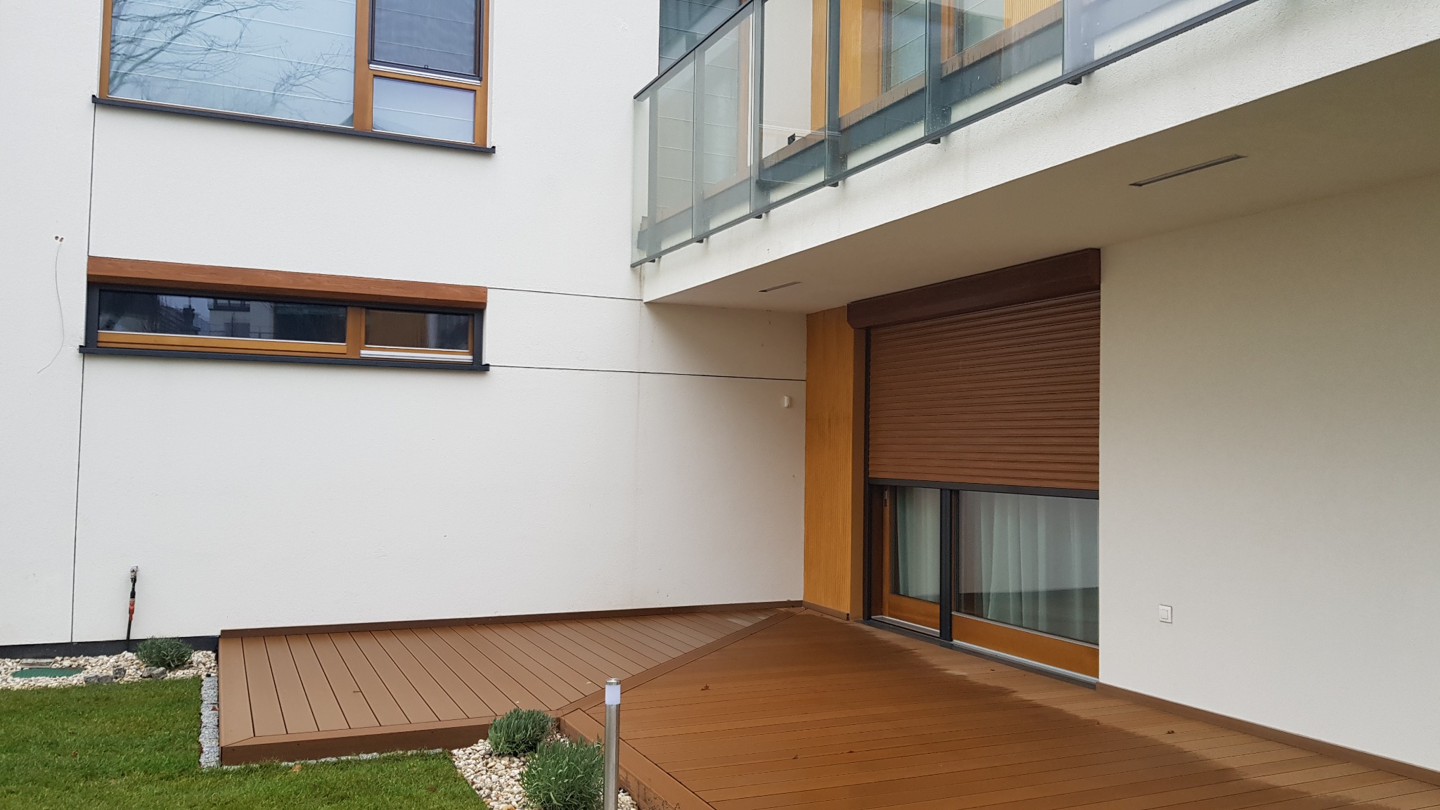 Brązowa roleta zewnętrzna opuszczona na oknie tarasowym w nowoczesnym domu w Otwocku, widok z poziomu tarasu z drewnianą podłogą.