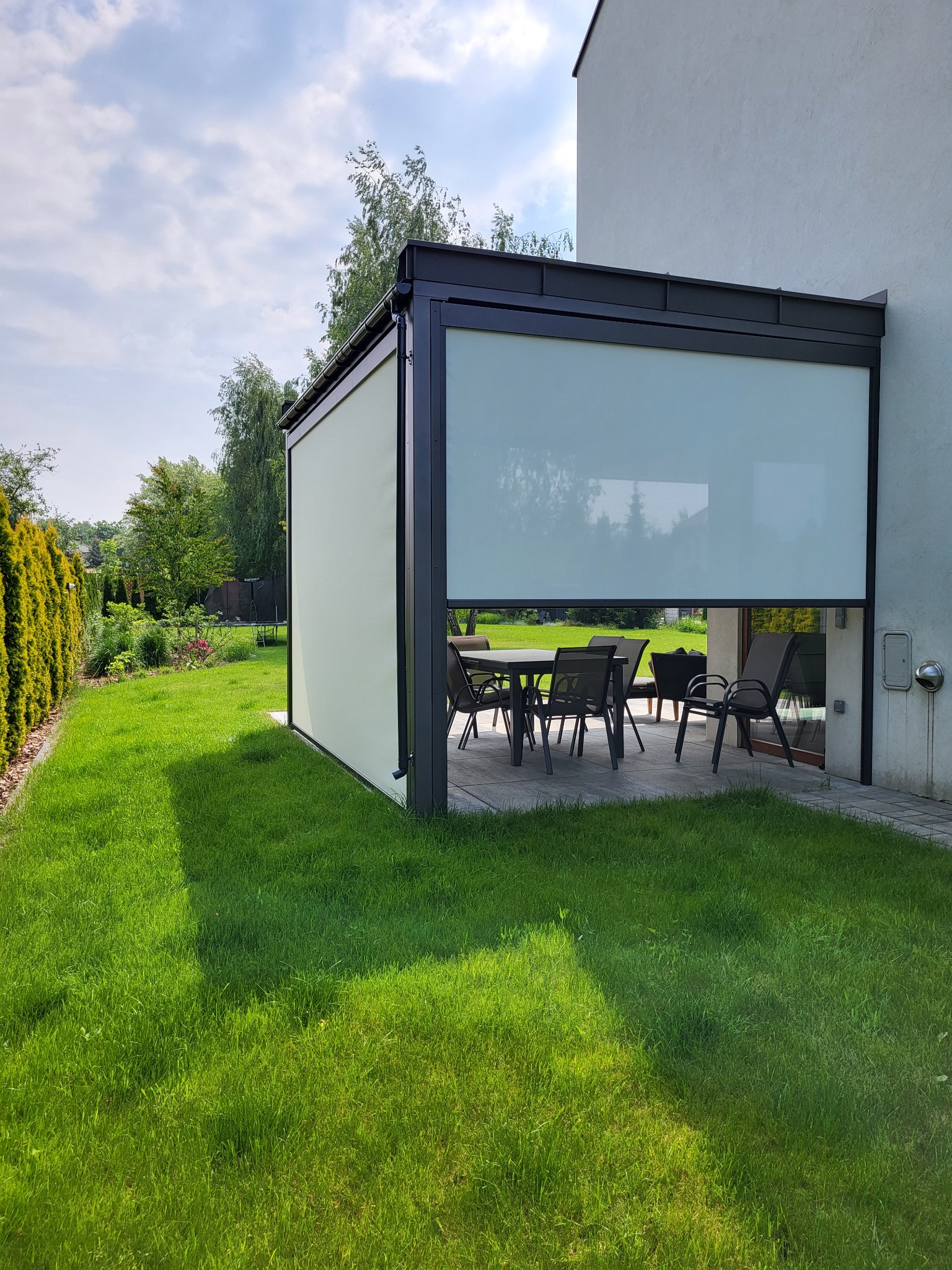 Zewnętrzna konstrukcja z opuszczoną, jasną przesłoną tekstylną, tworząca zacienione patio z meblami ogrodowymi, widok z trawnika w słoneczny dzień.