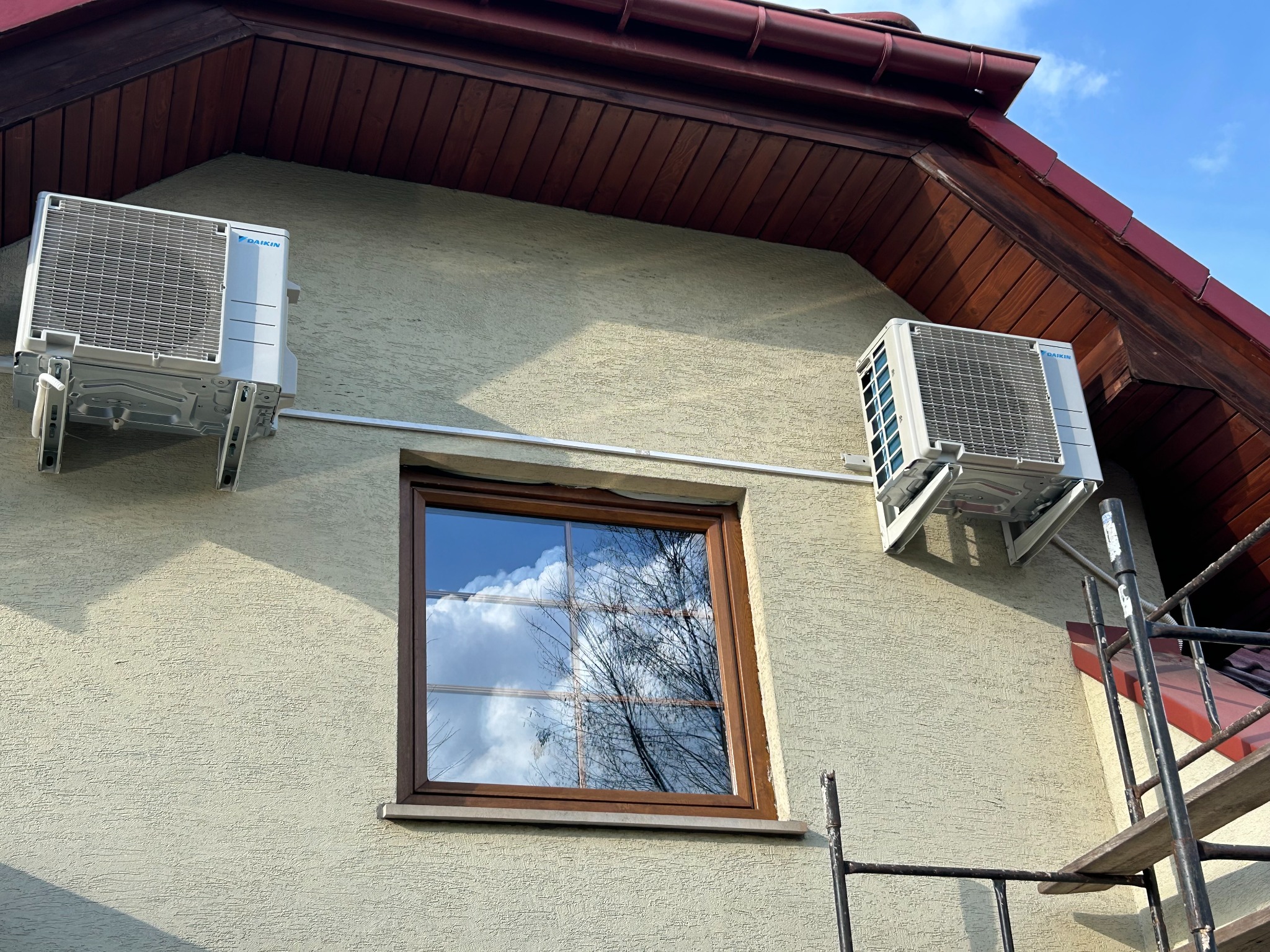 Dwa klimatyzatory Daikin zamontowane na elewacji domu z oknem, widoczne rusztowanie. Ujęcie z dołu, jasne niebo.