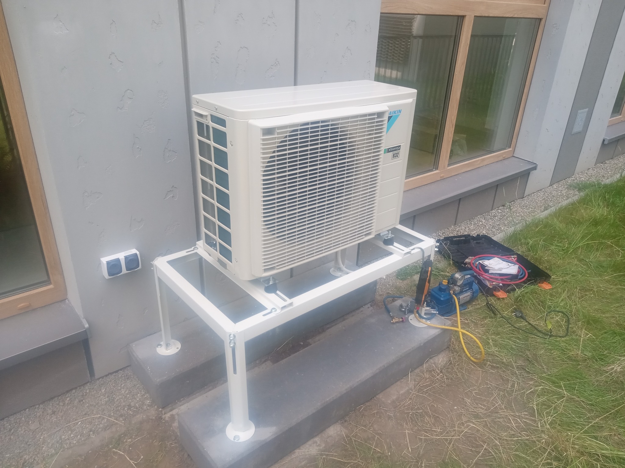 Biała jednostka zewnętrzna klimatyzacji Daikin na metalowym stelażu, zamontowana przy elewacji budynku z oknami. Obok widoczne narzędzia serwisowe.