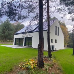 M.Konstruktywny Marek Krzyżyk - Nowoczesny, biały dom murowany z panelami słonecznymi na dachu, widoczny z perspektywy ogrodu, z drzewami na pierwszym planie i samochodem w tle.